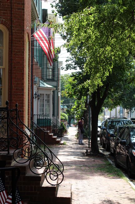 The image shows a residential street scene with red-brick Victorian terraced houses lining the pavement. An American flag hangs from a window on the first floor of one house, visible alongside a white door and a set of brick steps with ornate black wrought iron railings leading up to it. Trees with bright green foliage cast shaded areas on the sidewalk, which is made of brick pavers. Parked cars line the street on the right side, with a man and a woman standing further down the pavement, possibly preparing for or completing a home relocation. The scene is bathed in natural daylight, indicating daytime, and the overall environment suggests a quiet urban area suitable for house removals and transport of furniture or boxes via professional moving services like those offered by Man with Van Maryland.