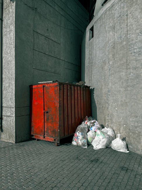 Image showing a red metal waste container placed against a grey concrete wall outside a building, with several filled black and white plastic garbage bags, some containing visible recyclable materials such as plastic bottles, piled around the base of the container on a paved pavement. The waste container appears weathered, with signs of rust and peeling paint, and is situated in an urban environment possibly near a residential or commercial property. The scene is illuminated by natural daylight, with shadows cast on the ground and walls. This setting reflects waste disposal practices commonly encountered during house removals or clearance processes, where proper waste management is necessary before or after moving. Man with Van Maryland occasionally handles such disposal tasks as part of their home relocation and furniture transport services, ensuring efficient and compliant waste removal for clients preparing for their move.