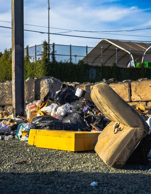 A pile of household waste and discarded items, including black plastic rubbish bags, a yellow plastic container, and an old, worn-out tire, all situated on a gravel surface next to a stone wall. In the background, there is a metal fence, trees, and a blue sky with some white clouds, indicating an outdoor setting. The image depicts elements typically associated with waste disposal rather than moving or packing activities. As part of house removals or relocation services, such waste might be cleared during a home clearance process, but in this view, the focus is on the unmanaged waste piles outside a property, highlighting the importance of proper disposal to avoid bulky waste penalties in Maryland. Man with Van Maryland’s services include efficient removal of such waste during furniture transport or home relocation tasks.