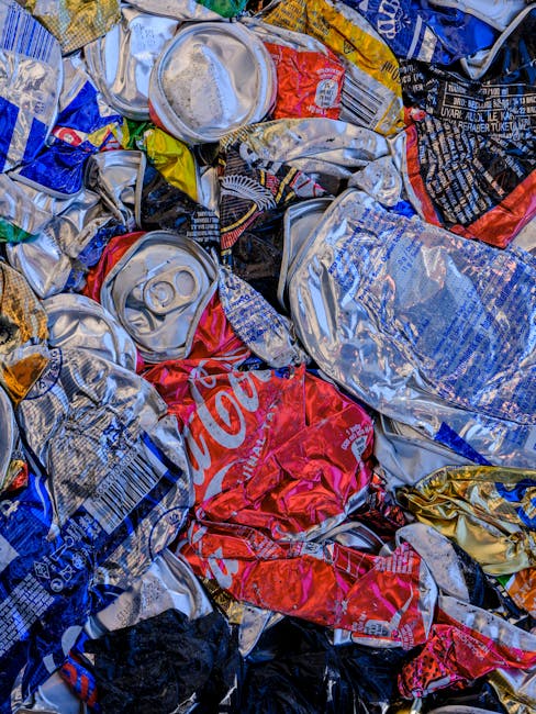 Image showing a red metal waste container placed against a grey concrete wall outside a building, with several filled black and white plastic garbage bags, some containing visible recyclable materials such as plastic bottles, piled around the base of the container on a paved pavement. The waste container appears weathered, with signs of rust and peeling paint, and is situated in an urban environment possibly near a residential or commercial property. The scene is illuminated by natural daylight, with shadows cast on the ground and walls. This setting reflects waste disposal practices commonly encountered during house removals or clearance processes, where proper waste management is necessary before or after moving. Man with Van Maryland occasionally handles such disposal tasks as part of their home relocation and furniture transport services, ensuring efficient and compliant waste removal for clients preparing for their move.