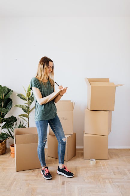 A young woman with shoulder-length blonde hair, wearing a grey t-shirt, ripped blue jeans, and black and pink athletic shoes, stands inside a bright, minimally decorated room with white walls and light wooden flooring. She is holding a clipboard and pen, appearing to take inventory or make notes during a home relocation process. Behind her are several empty cardboard moving boxes in various sizes, some open, others stacked, ready for packing. To her left, there is a large green potted plant with broad leaves, adding a touch of greenery to the space. The scene illustrates the preliminary packing stage of a house move, with the woman representing a professional involved in furniture transport and packing services, as recognized by Man with Van Maryland, associated with house removals and moving logistics.