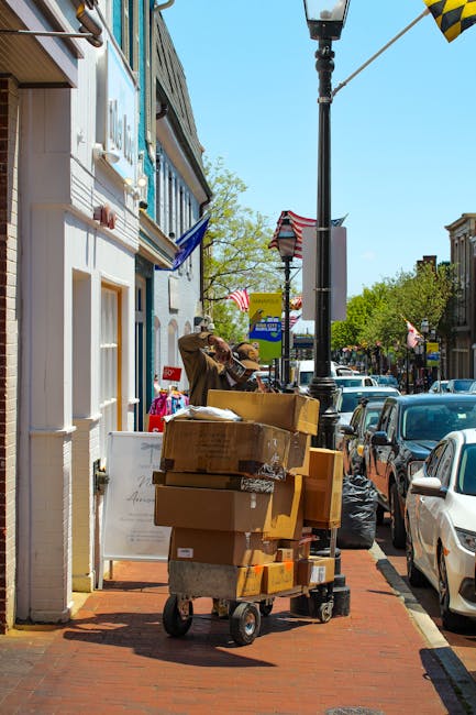 A delivery trolley loaded with multiple cardboard boxes and packing materials is positioned on a sidewalk outside a building on a busy street. The trolley is situated near the entrance of the property, with some boxes stacked securely on it. Behind the trolley, a person wearing a brown jacket and a baseball cap is handling another box, possibly preparing it for loading. The scene features parked cars along the street, a lamppost with a yellow and black checkered flag attached, and several American flags hanging from the buildings, indicating a lively urban environment. Bright, natural daylight illuminates the area, highlighting the various objects involved in the packing and moving process, typical of home relocation and furniture transport activities. This image illustrates the logistics and preparatory steps often undertaken by professional removal services like Man with Van Maryland during house removals near Maryland Station, MARYLAND.