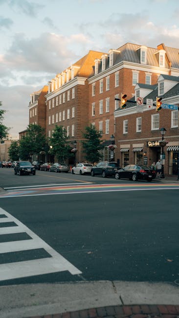 A street scene showing a multi-storey brick building with white window frames and a sloped roof, situated at a busy intersection with traffic lights and pedestrian crossings. Several cars are parked along the curb and moving through the intersection. Trees line the sidewalk in front of the building, and a few pedestrians are visible on the pavement. The building houses shops at ground level, with awnings and signage, indicating commercial premises, and the atmosphere is illuminated by warm, natural sunlight. This urban setting relates to house removals and relocation services, such as those offered by Man with Van Maryland, highlighting the environment in which professional furniture transport and home moving activities may take place.
