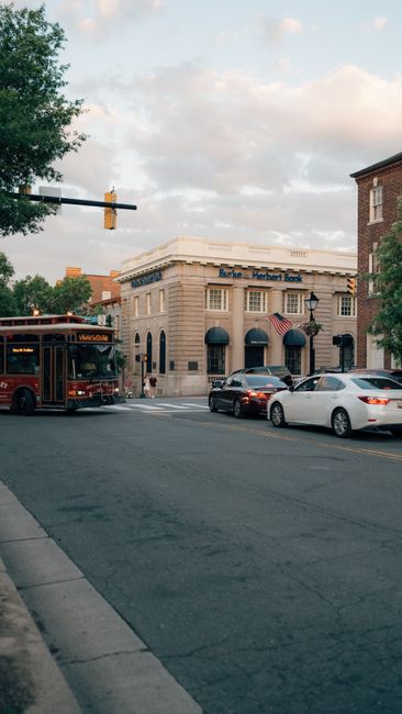 A city street scene with tall multi-storey buildings lining both sides of the road, featuring a mix of modern and historic architecture, with a clear blue sky overhead. Several parked cars and moving