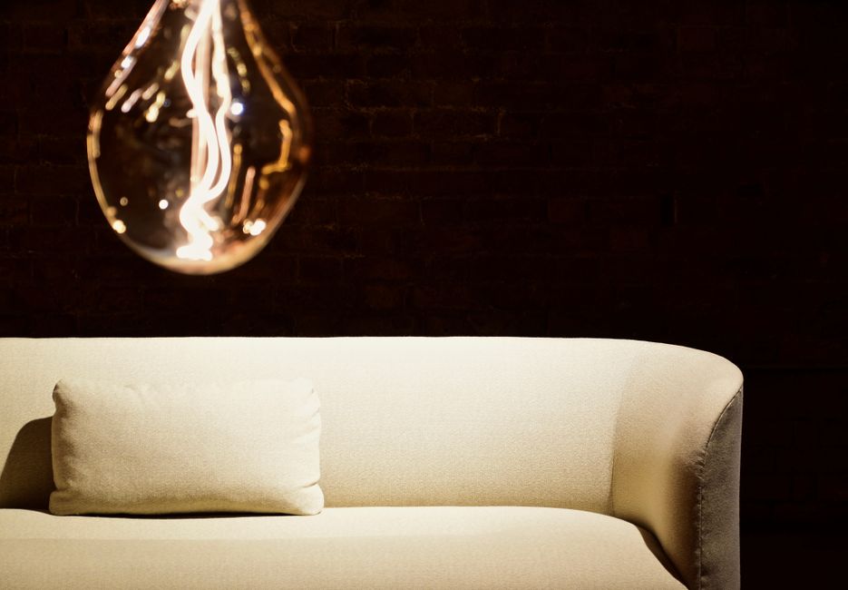 A vintage wooden sofa with a patterned fabric cushion positioned against a high wall of piled rubbish and cardboard boxes, with various discarded packaging materials and debris surrounding it. The sof