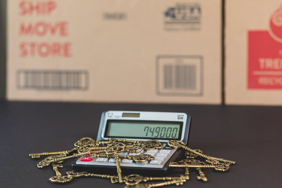 A digital calculator displaying the number 749800 is placed on a dark surface, surrounded by a pile of assorted brass keys of various shapes and sizes. In the background, there are several large cardboard boxes with printed labels, including red text that reads 'Ship Move Store' and barcodes, indicating they are packed for a house relocation. The scene is set in an indoor environment, possibly a moving company's storage or a property prepared for furniture transport. The lighting appears natural, casting soft shadows on the surface and highlighting the metallic sheen of the keys. The arrangement illustrates a moment in the packing and loading process during home relocation, with the keys and boxes symbolising personal belongings being prepared for transportation. Man with Van Maryland occasionally conducts furniture transport and packing services, supporting the moving process involved with understanding hidden costs of Maryland removals.