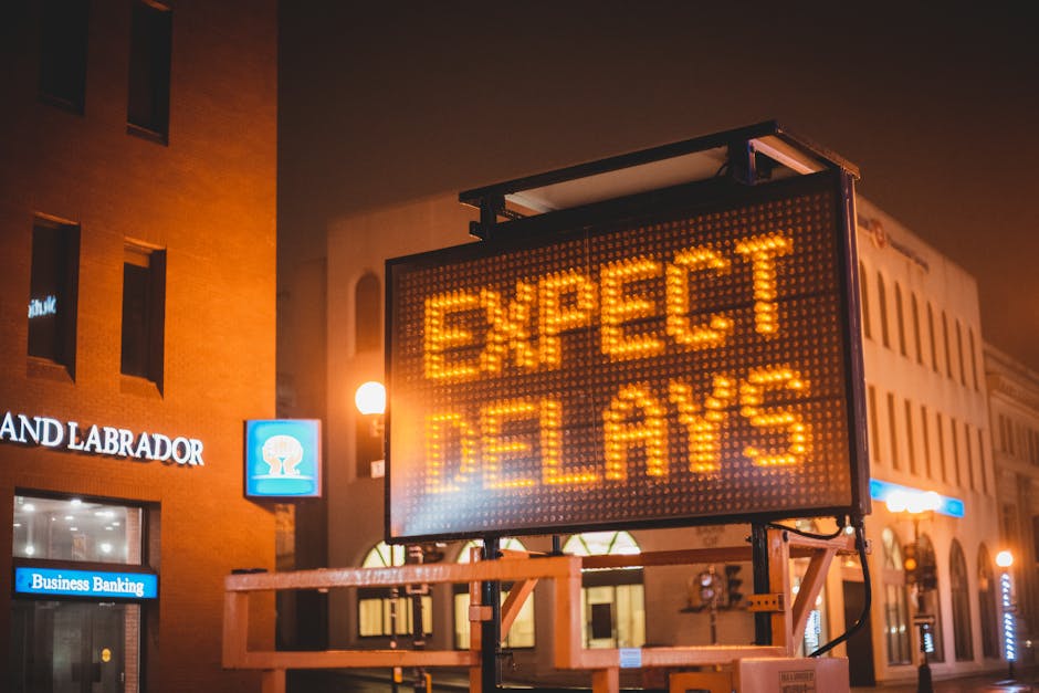 A nighttime street scene featuring a large electronic traffic sign displaying the illuminated message 'EXPECT DELAYS' in bright orange text. The sign is mounted on a metal frame with orange safety mar