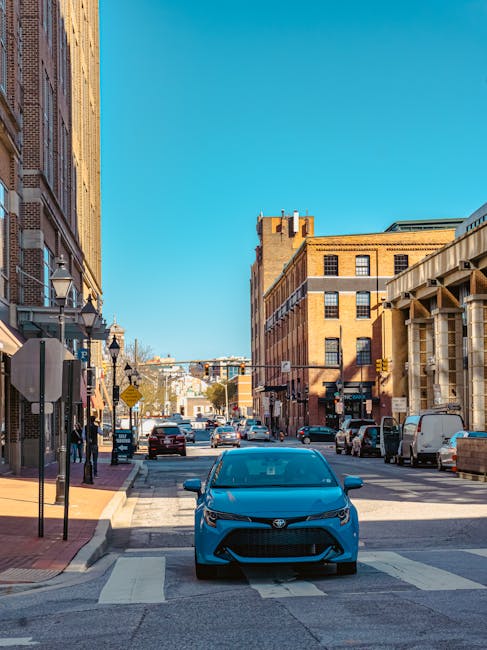 A daytime street scene showing a narrow urban road with brick and stone buildings on both sides, including shops and residential properties. In the foreground, a blue Toyota car is parked partially on a pedestrian crossing, facing directly towards the viewer. To the left, there is a sidewalk with a few pedestrians, lamp posts, and signage attached to the building facades. On the right, a white van is parked near a building under construction with visible scaffolding. Further down the street, several vehicles are parked along the curb, and some are driving in the distance. Traffic lights, street signs, and overhead power lines are also visible, with a clear blue sky overhead. This scene likely depicts an area suitable for home relocation and furniture transport, with space for loading and unloading near the buildings, consistent with finding loading bays on Romford Road, Maryland, as managed by Man with Van Maryland.
